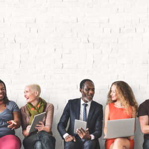 Blog Default Image. Group of people sitting against a wall sharing things on electronic devices.